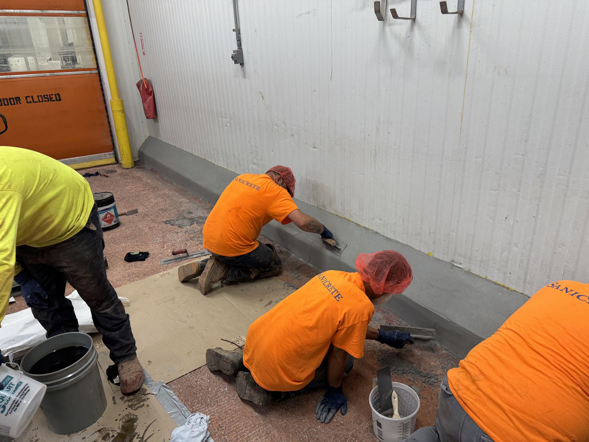 SaniCrete crew in orange shirts troweling VR 2-inch cant cove curbing along wall with orange bay door