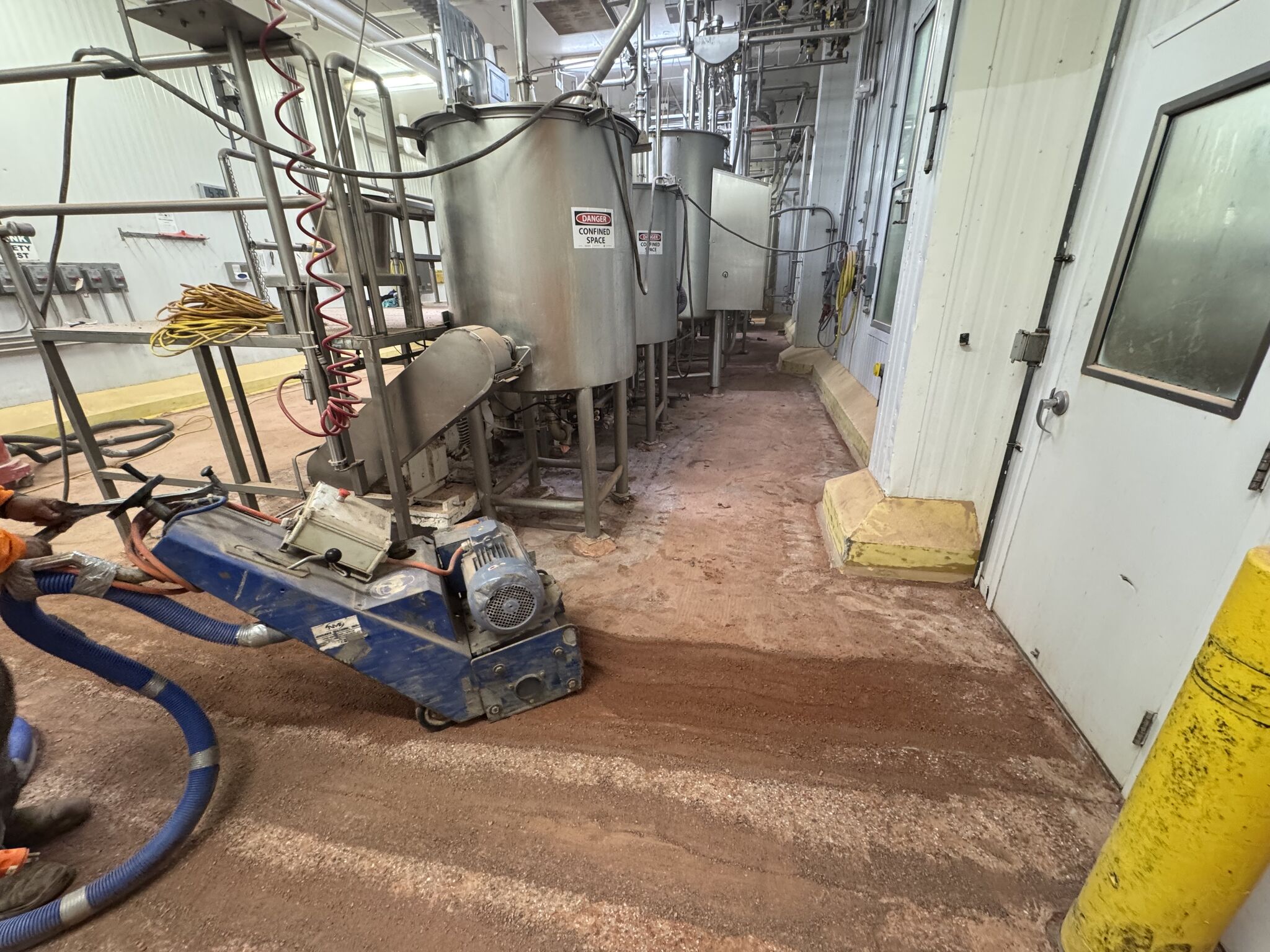 Crew member operating blue scarifier to profile concrete substrate around stainless tanks and yellow bollards
